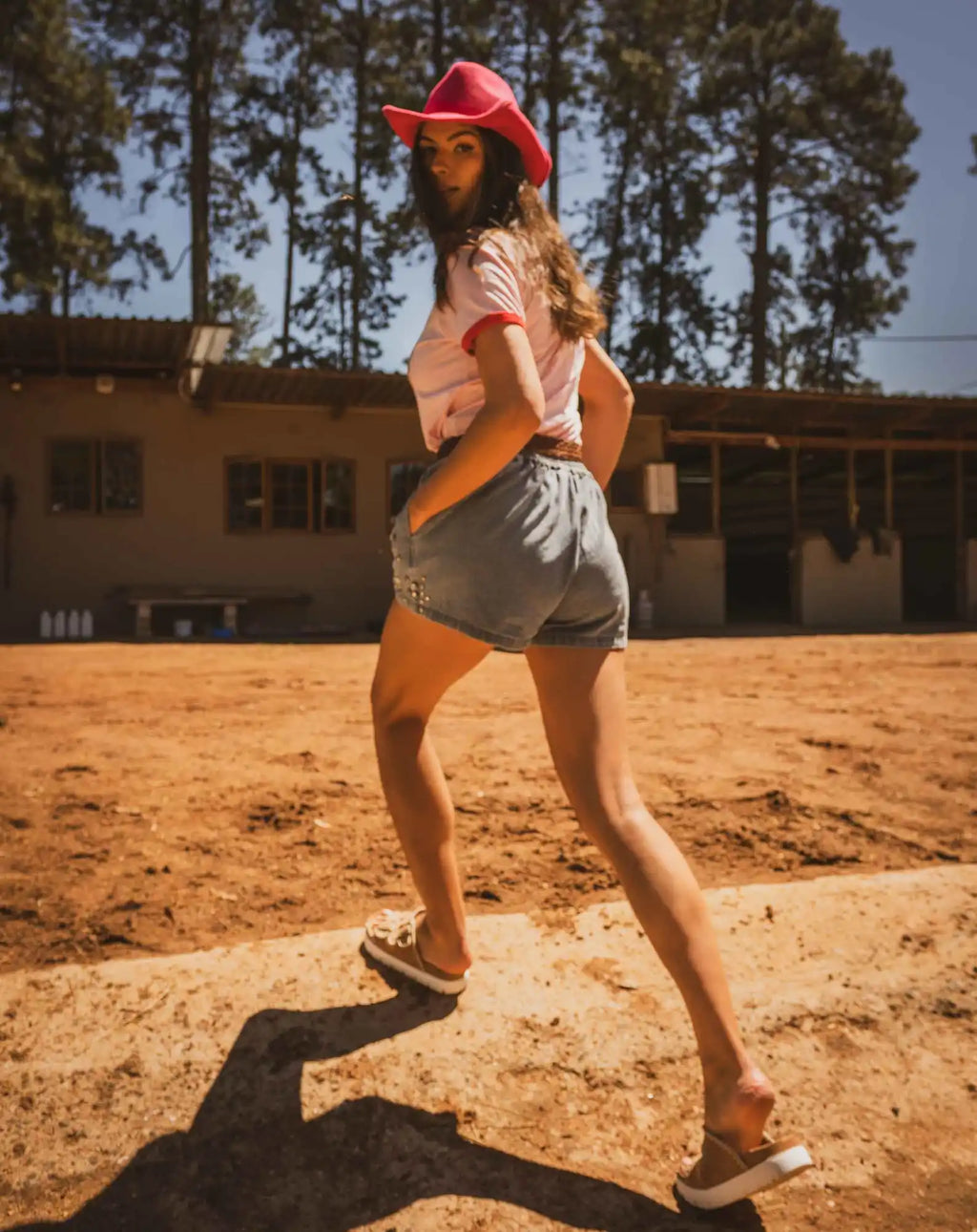 A person wearing a red baseball cap, pink shirt and gray shorts poses playfully on dusty ground.